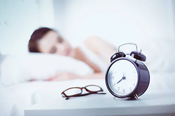 Rahat yatakta yatarken ve blissfully çalar saat arka plan üzerinde uyuyan güzel genç kadın — Stok fotoğraf