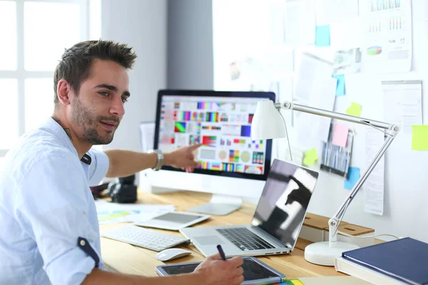 Retrato del joven diseñador sentado en el estudio gráfico frente a la computadora portátil y el ordenador mientras trabaja en línea. — Foto de Stock