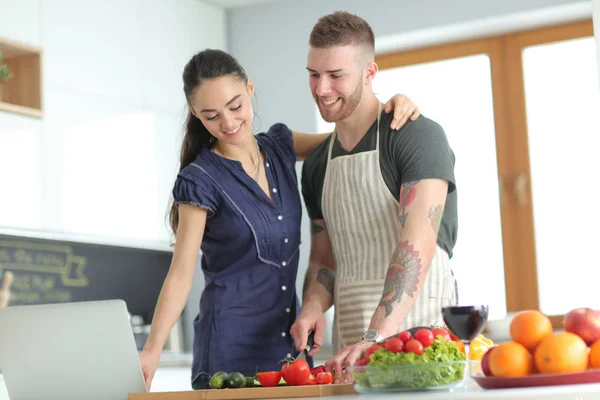 Junger Mann schneidet Gemüse und Frau steht mit Laptop in der Küche — Stockfoto