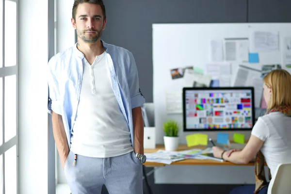 Porträt eines jungen Designers vor Laptop und Computer während der Arbeit. Assistentin bedient ihr Handy im Hintergrund. — Stockfoto