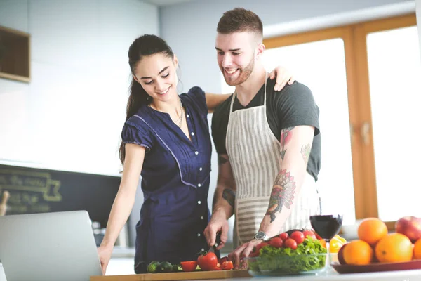 Ung man skär grönsaker och kvinna stående med laptop i köket — Stockfoto