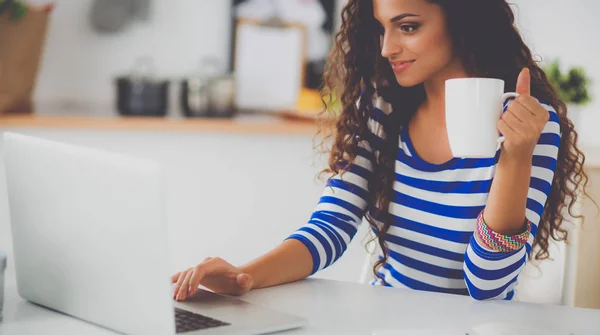 Mujer joven sonriente con taza de café y portátil en la cocina en casa — Foto de Stock