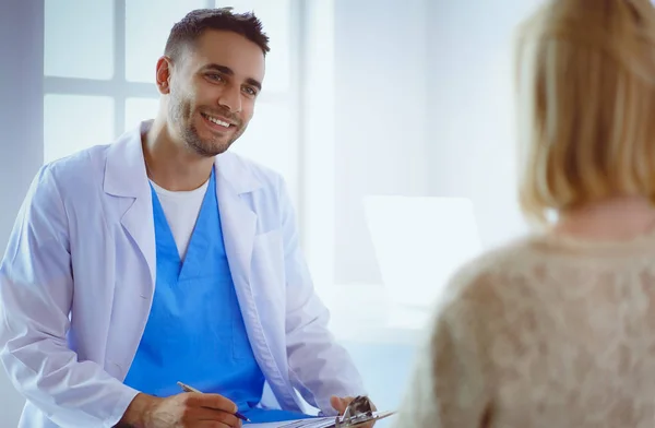 Schöner Arzt spricht mit junger Patientin und macht sich Notizen, während er in seinem Büro sitzt — Stockfoto