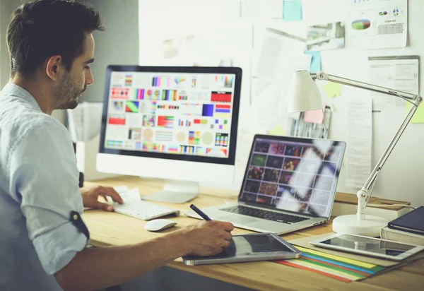 Retrato del joven diseñador sentado en el estudio gráfico frente a la computadora portátil y el ordenador mientras trabaja en línea — Foto de Stock