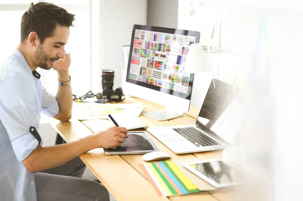 Retrato del joven diseñador sentado en el estudio gráfico frente a la computadora portátil y el ordenador mientras trabaja en línea — Foto de Stock