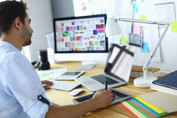 Retrato del joven diseñador sentado en el estudio gráfico frente a la computadora portátil y el ordenador mientras trabaja en línea — Foto de Stock