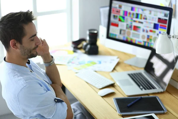 Retrato del joven diseñador sentado en el estudio gráfico frente a la computadora portátil y el ordenador mientras trabaja en línea — Foto de Stock