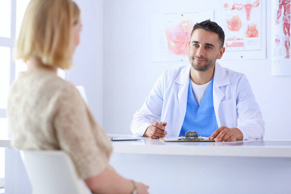 Красивый врач разговаривает с молодой пациенткой и делает заметки, сидя в своем кабинете