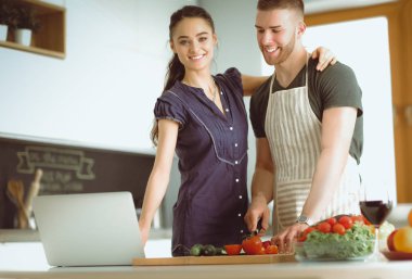 Sebze doğrayan genç bir adam ve mutfakta dizüstü bilgisayarı olan bir kadın.