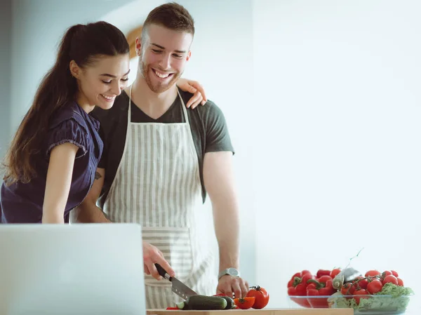Hombre joven cortando verduras y mujer de pie con el ordenador portátil en la cocina — Foto de Stock