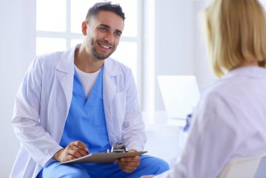 Yakışıklı doktor genç bayan hastasıyla konuşuyor ve ofisinde otururken notlar alıyor.