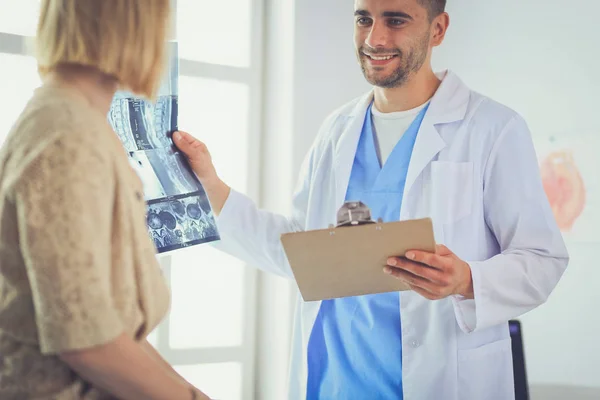 Knappe dokter praat met jonge vrouwelijke patiënt en maakt aantekeningen terwijl hij in zijn kantoor zit — Stockfoto