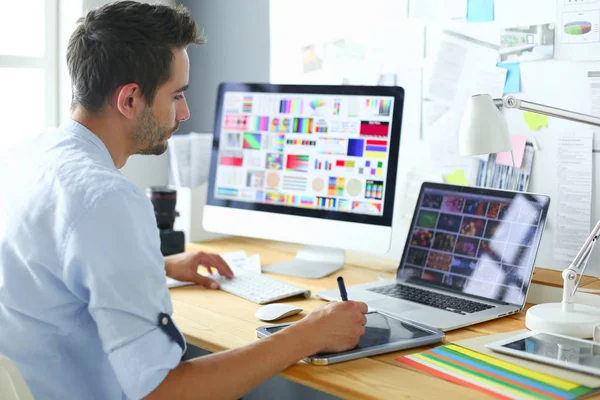 Retrato del joven diseñador sentado en el estudio gráfico frente a la computadora portátil y el ordenador mientras trabaja en línea. — Foto de Stock