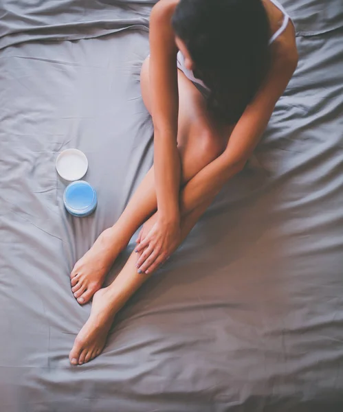 Beautiful woman sitting on bed and applying cream on legs — Stock Photo, Image