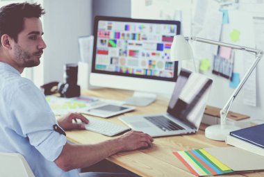 İnternette çalışırken bilgisayarın ve dizüstü bilgisayarın önünde grafik stüdyosunda oturan genç tasarımcının portresi.