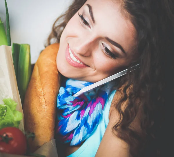 Smiling woman with mobile phone holding shopping bag in kitchen — Stock Photo, Image