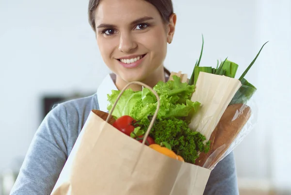 Mujer joven sosteniendo bolsa de la compra de comestibles con verduras de pie en la cocina. —  Fotos de Stock