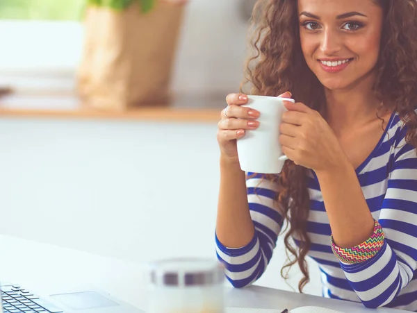 Smiling young woman with coffee cup and laptop in the kitchen at home — Stock Photo, Image