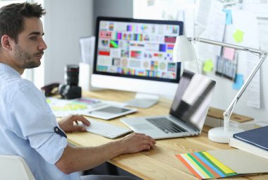 İnternette çalışırken bilgisayarın ve dizüstü bilgisayarın önünde grafik stüdyosunda oturan genç tasarımcının portresi.