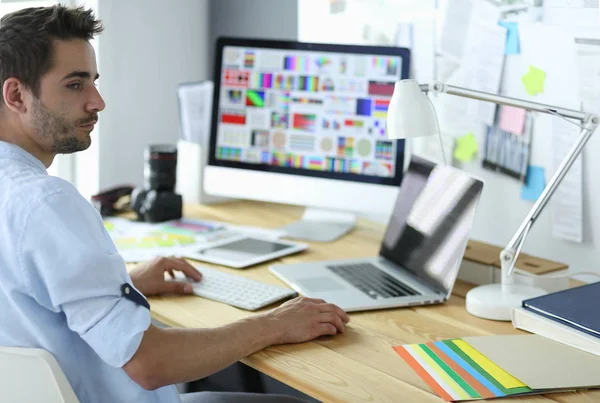 Retrato del joven diseñador sentado en el estudio gráfico frente a la computadora portátil y el ordenador mientras trabaja en línea. — Foto de Stock