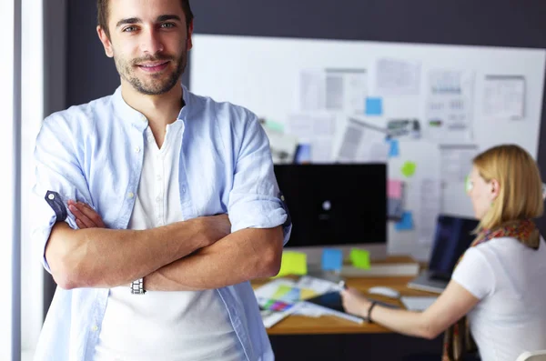 Porträt eines jungen Designers vor Laptop und Computer während der Arbeit. Assistentin bedient ihr Handy im Hintergrund. — Stockfoto