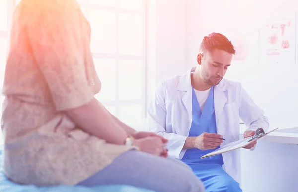 El guapo doctor está hablando con una paciente joven y tomando notas mientras está sentado en su consultorio.. —  Fotos de Stock