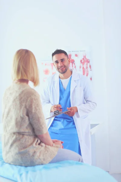 Knappe dokter praat met jonge vrouwelijke patiënt en maakt aantekeningen terwijl hij in zijn kantoor zit. — Stockfoto