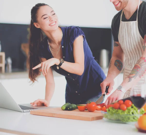 Hombre joven cortando verduras y mujer de pie con el ordenador portátil en la cocina — Foto de Stock