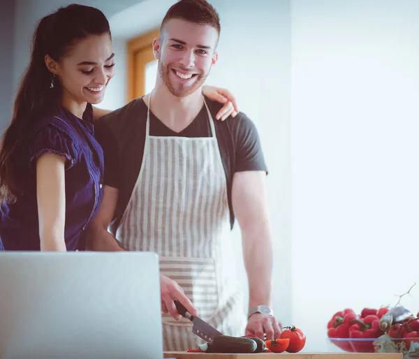 Junger Mann schneidet Gemüse und Frau steht mit Laptop in der Küche — Stockfoto