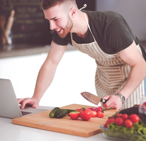 Portrait of man cooking vegetable in the kitchen while looking at a laptop computer on the table — Stock Photo, Image