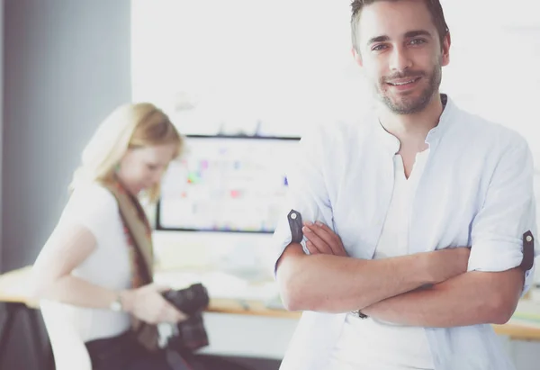 Porträt eines jungen Designers vor Laptop und Computer während der Arbeit. Assistentin bedient ihr Handy im Hintergrund. — Stockfoto