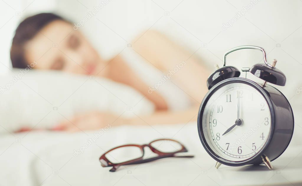 Beautiful young woman sleeping while lying in bed comfortably and blissfully on the background of alarm clock