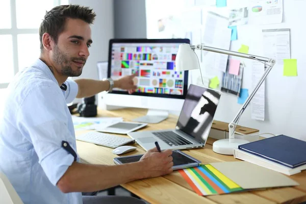 Retrato del joven diseñador sentado en el estudio gráfico frente a la computadora portátil y el ordenador mientras trabaja en línea. — Foto de Stock