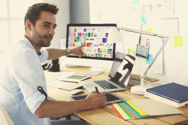 Retrato del joven diseñador sentado en el estudio gráfico frente a la computadora portátil y el ordenador mientras trabaja en línea. — Foto de Stock