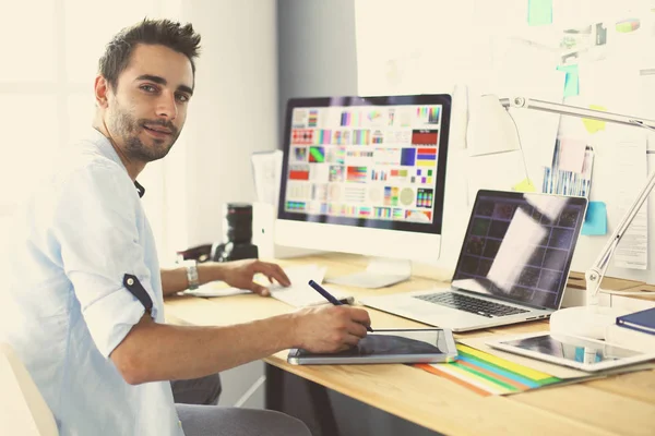 Retrato del joven diseñador sentado en el estudio gráfico frente a la computadora portátil y el ordenador mientras trabaja en línea. — Foto de Stock