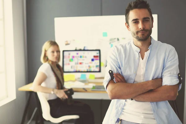 Porträt eines jungen Designers vor Laptop und Computer während der Arbeit. Assistentin bedient ihr Handy im Hintergrund. — Stockfoto