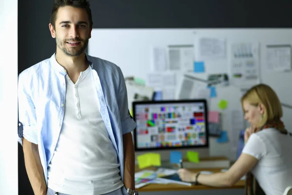 Porträt eines jungen Designers vor Laptop und Computer während der Arbeit. Assistentin bedient ihr Handy im Hintergrund. — Stockfoto