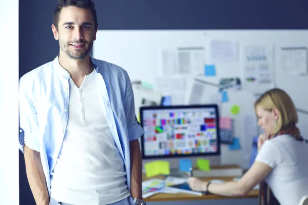 Retrato del joven diseñador delante de la computadora portátil y el ordenador mientras trabaja. Asistente usando su móvil en segundo plano . — Foto de Stock