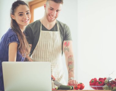 Sebze doğrayan genç bir adam ve mutfakta dizüstü bilgisayarı olan bir kadın.