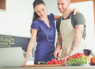 Sebze doğrayan genç bir adam ve mutfakta dizüstü bilgisayarı olan bir kadın.