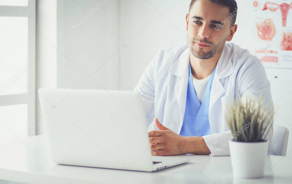 Portrait of a male doctor with laptop sitting at desk in medical office.