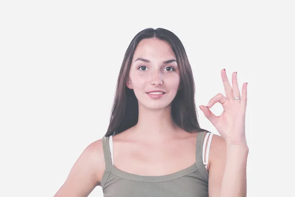 Beautiful young woman is looking at camera, showing Ok sign and smiling, standing against gray wall — Stock Photo, Image