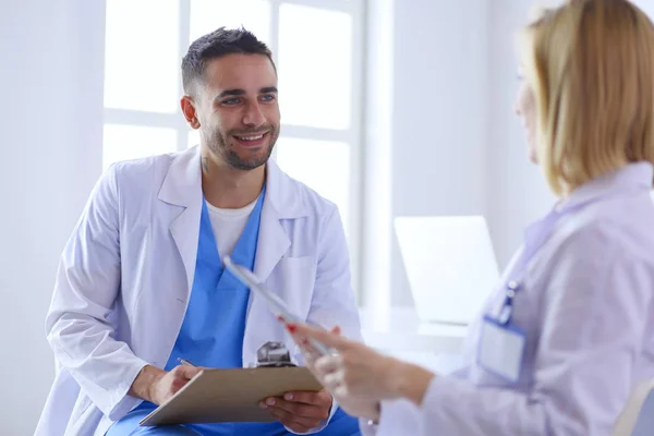 Schöner Arzt spricht mit junger Ärztin und macht sich Notizen, während er in seinem Büro sitzt. — Stockfoto