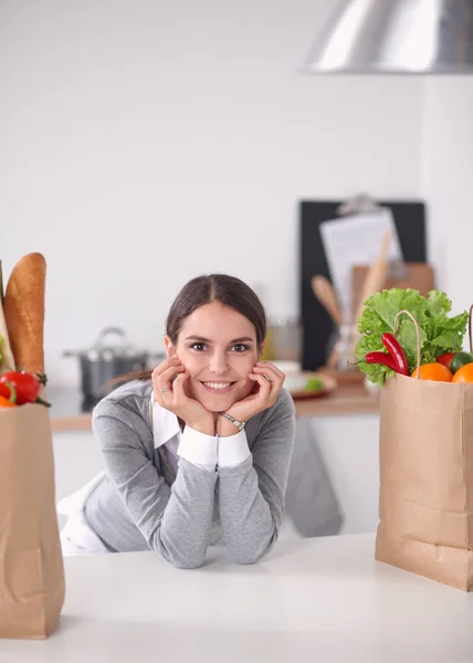 Mujer joven sosteniendo bolsa de la compra de comestibles con verduras de pie en la cocina —  Fotos de Stock