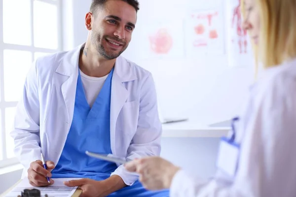 Schöner Arzt spricht mit junger Ärztin und macht sich Notizen, während er in seinem Büro sitzt. — Stockfoto