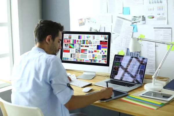 Retrato del joven diseñador sentado en el estudio gráfico frente a la computadora portátil y el ordenador mientras trabaja en línea. — Foto de Stock