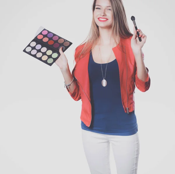 Ung vacker kvinna håller i ena handen och en palett med färg och skuggor för makeup, begagnad pensel för makeup. — Stockfoto
