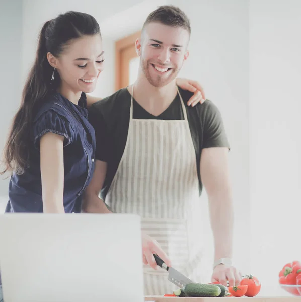 Hombre joven cortando verduras y mujer de pie con el ordenador portátil en la cocina — Foto de Stock