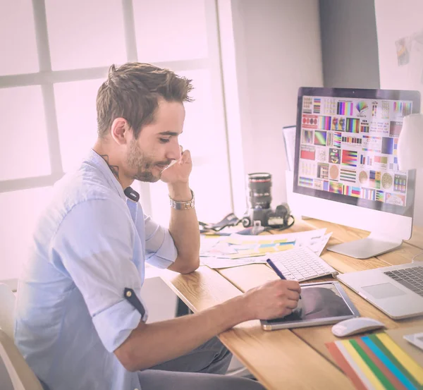 Retrato del joven diseñador sentado en el estudio gráfico frente a la computadora portátil y el ordenador mientras trabaja en línea. — Foto de Stock