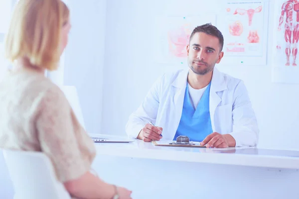 Bello medico sta parlando con giovane paziente femminile e prendere appunti mentre seduto nel suo ufficio. — Foto Stock